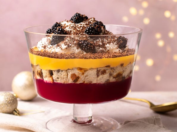 A layered trifle in a glass dish with a red jelly base, sponge pieces, yellow custard, whipped cream, blackberries, and chocolate shavings, set on a light surface with festive decorations in the background.