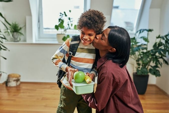 A woman kisses her smiling son on the cheek as she hands him a container with an apple and a banana, a simple healthy school lunchbox idea.