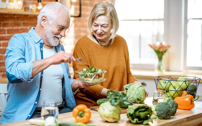 An older man and woman smile together in a bright kitchen while preparing a fresh salad. The table is covered with vegetables like cabbage, pepper, and broccoli, along with a bowl of apples and glasses of water.