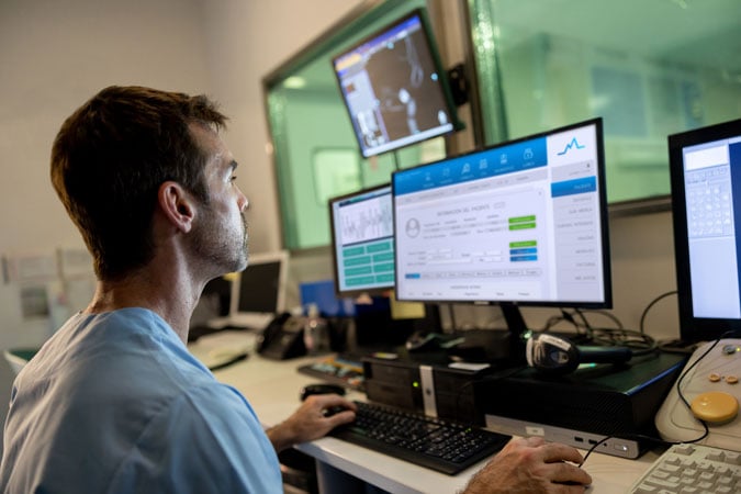 A healthcare professional in scrubs sits at a desk, reviewing medical data on monitors in a clinical control room, monitoring patients living with bowel cancer.