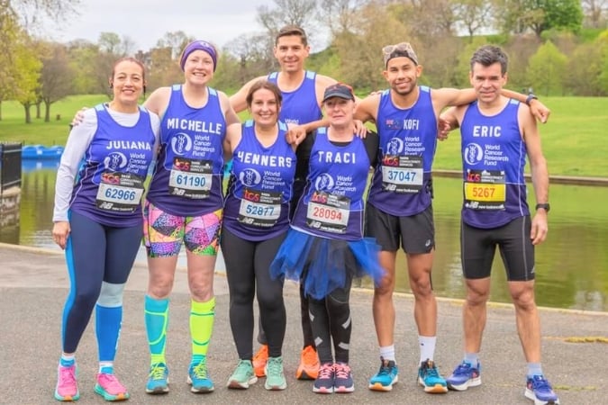 Seven smiling marathon runners in matching blue shirts pose together outdoors near a park lake. Each wears a race number with their names: Juliana, Michelle, Henners, Traci, Kofi, and Eric. It is a cloudy day.