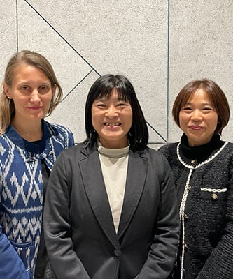 Dr Sarah Abe, Dr Manami Inoue and Dr Norie Sawada are standing together indoors, smiling at the camera. They are wearing jumpers and jackets, and the background has a light-coloured wall with geometric lines.