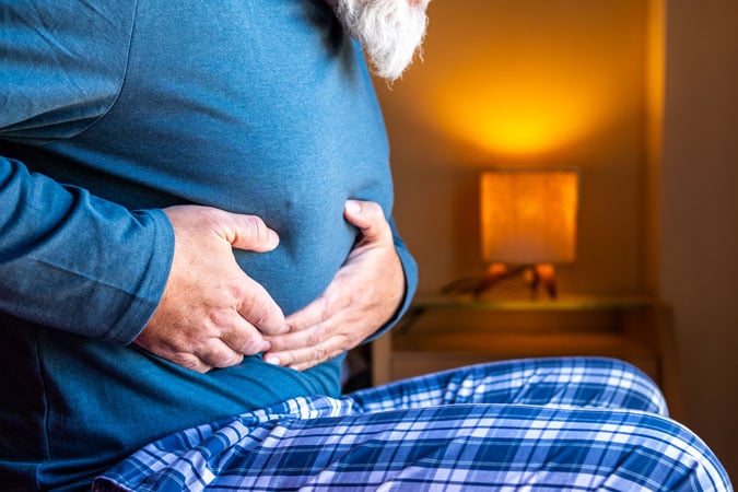 A person living with bowel cancer, wearing a blue long-sleeved shirt and checked pyjama bottoms, sits holding their abdomen in discomfort. A lamp glows softly in the background.