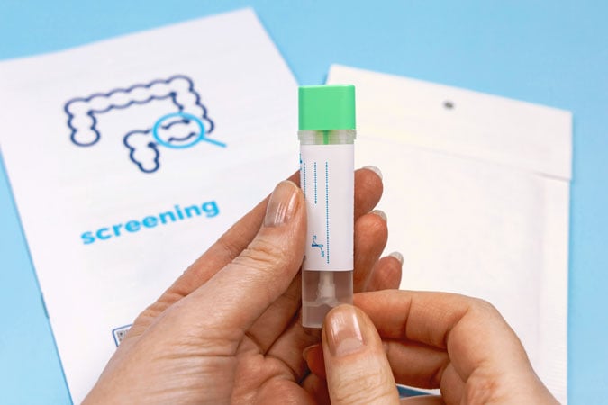 A person holds a sample collection tube with a green cap. In the background, there is a screening kit envelope and an illustrated instruction sheet showing a diagram of the colon—important tools for those living with bowel cancer.