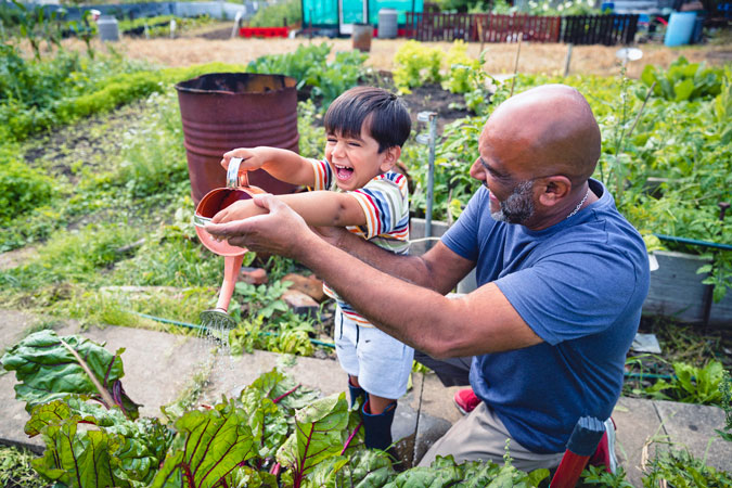 A smiling man helps a laughing young boy water plants with a small watering can in a lush vegetable garden on a sunny day.
