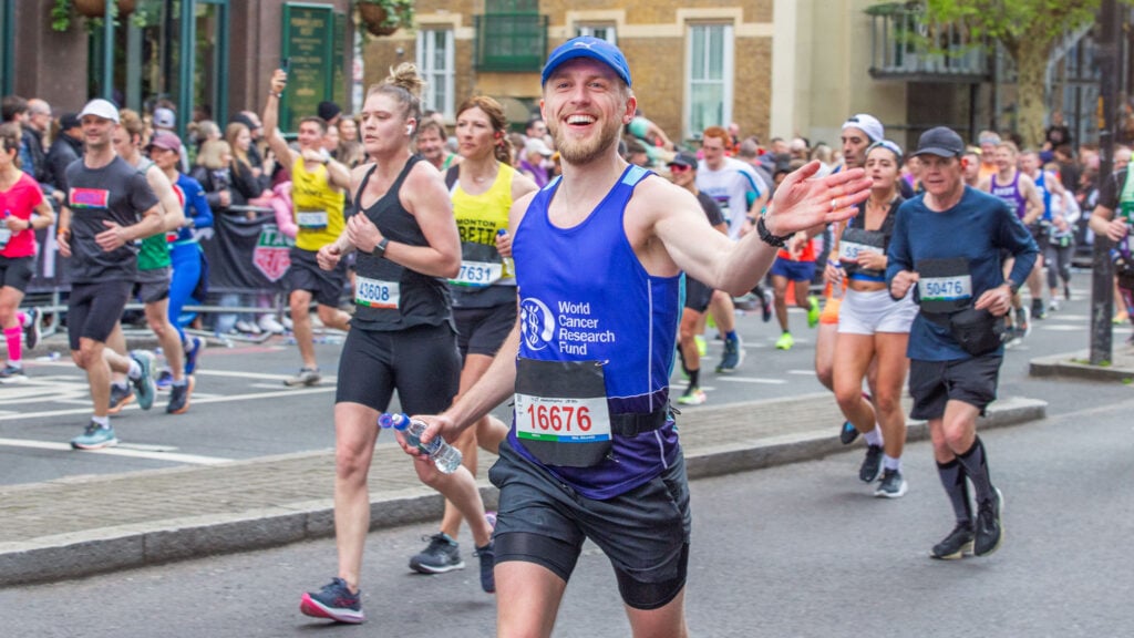 A smiling marathon runner wearing a blue cap and a World Cancer Research Fund vest waves at the camera whilst holding a water bottle, surrounded by other runners on a city street.