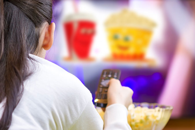 Teenager girl with remote control laying down and watching tv eating popcorn.