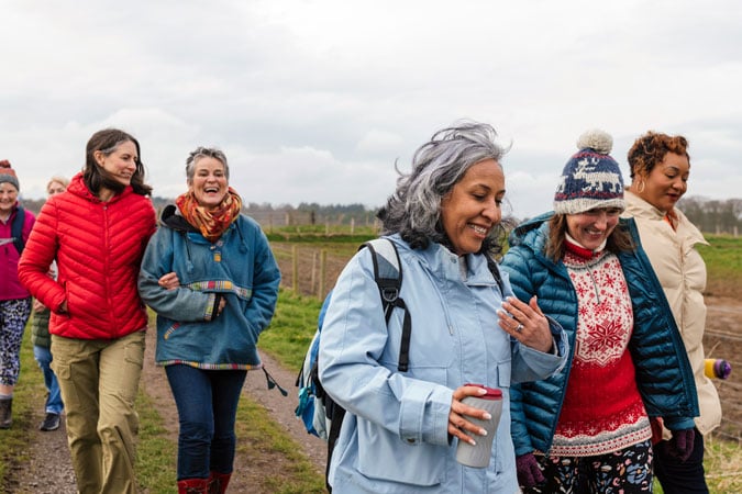 A group of women dressed in colourful winter clothing walk and laugh together outdoors on a cloudy day, with grassy fields and trees in the background.