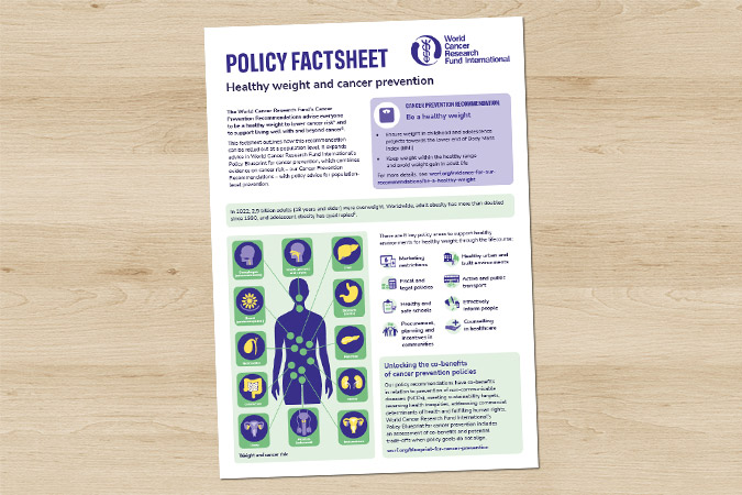 A fact sheet titled Healthy weight and cancer prevention policy with text, infographics of a human body, icons of organs, and health tips, placed on a wooden surface.