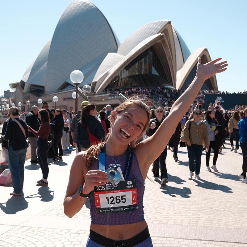 A smiling female runner holds her medal and raises one arm in celebration in front of the Sydney Opera House, surrounded by a crowd of people.