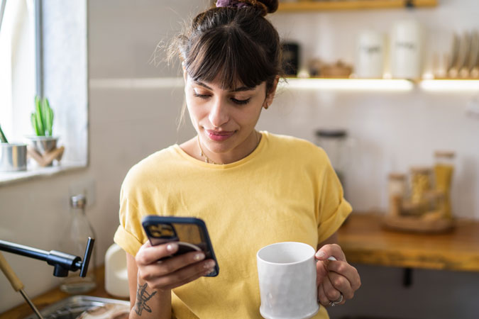 Young woman using the mobile phone while drinking coffee or tea at home