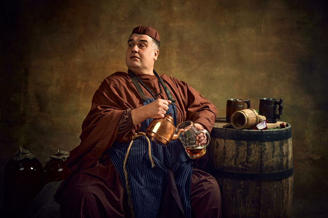 Medieval monk in period clothes pouring beer into mug from kettle, sitting in dimly lit cellar with wooden barrel and beer mug on top. Concept of comparison of eras, brewery, traditions (Getty Images)