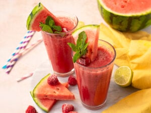 Two glasses of watermelon smoothie with mint, raspberries, and watermelon slices, surrounded by fresh fruit, straws, and a yellow cloth on a light surface.