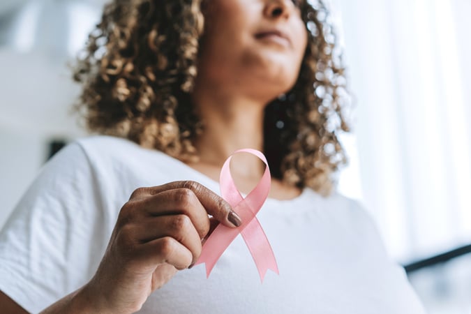 A woman with curly hair, wearing a white shirt, holds a pink breast cancer awareness ribbon in her hand. The focus is on the ribbon, symbolising support for breast cancer awareness.