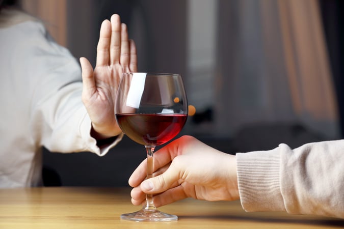 A person holds up their hand in a stop gesture whilst another person offers them a glass of red wine, indicating refusal of the drink.