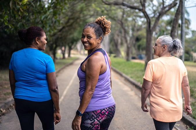 Three older women walk together on a tree-lined path. Two face forward whilst the woman in the centre, wearing a purple top, looks back and smiles at the camera. They appear to be enjoying a walk outdoors.