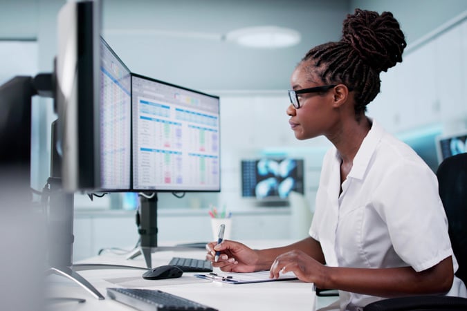 A woman wearing glasses and a white lab coat works at a desk with large computer monitors displaying data charts. She writes in a notebook whilst focussing on the screen in a modern office setting.