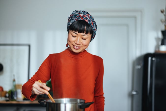 A woman wearing a patterned headscarf and an orange top smiles whilst stirring food in a pot in a kitchen.
