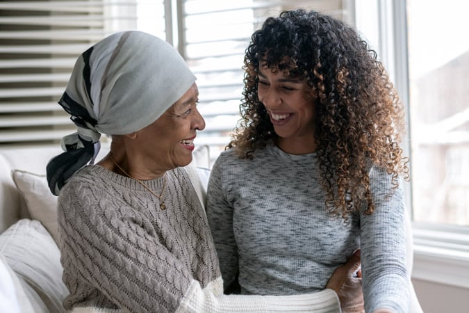 An older woman wearing a headscarf and a younger woman with curly hair sit close together on a sofa, smiling warmly at each other in a sunlit room.