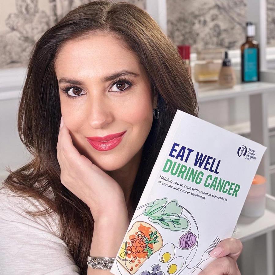 A woman with long brown hair smiles and holds a book titled Eat Well During Cancer with a colourful food illustration on the cover. She is indoors, with skincare bottles visible in the background.