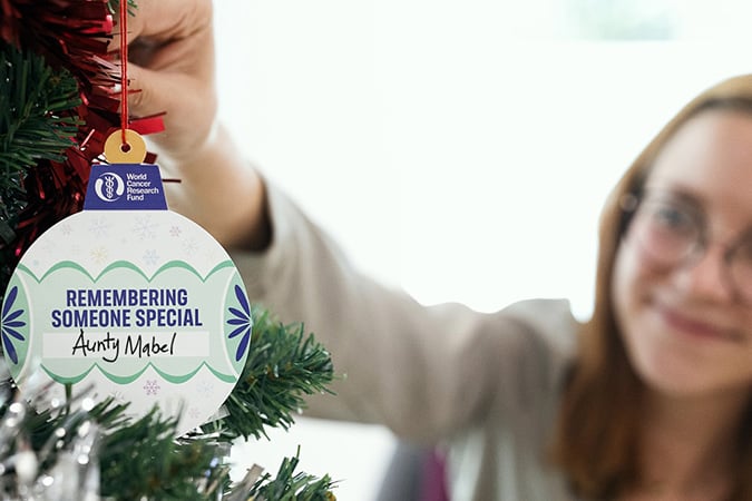 A person hangs a remembrance bauble on a Christmas tree. The bauble reads Remembering someone special, Auntie Mabel and features the World Cancer Research Fund logo. The person is smiling softly in the background.