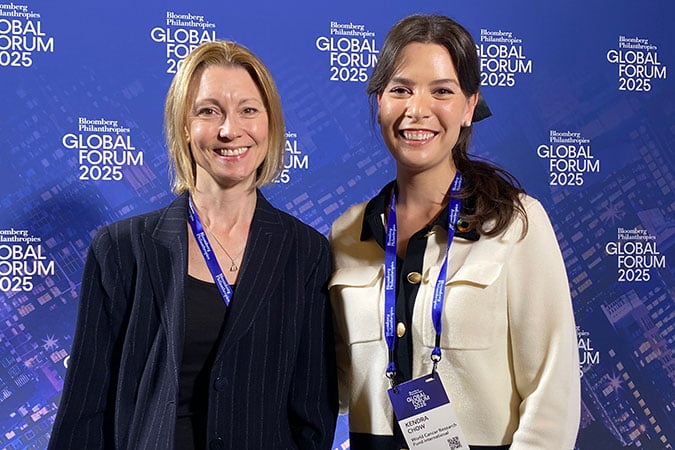 Dr Helen Croker and Kendra Chow standing smiling in front of a blue backdrop with Bloomberg Philanthropies GLOBAL FORUM 2025 repeatedly printed in white text.