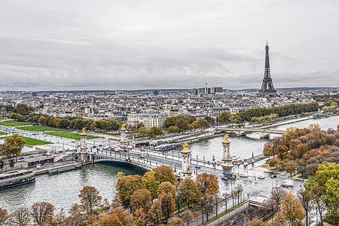 A panoramic view of Paris featuring the Eiffel Tower, the Seine River, and the ornate Pont Alexandre III bridge surrounded by trees with autumn foliage under a cloudy sky.