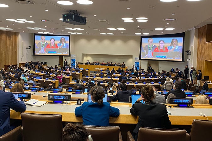 Inside the UN High-Level Meeting for NCDs: A large conference room filled with people listening to speakers at a panel discussion; two screens display three women presenting at the front of the room.