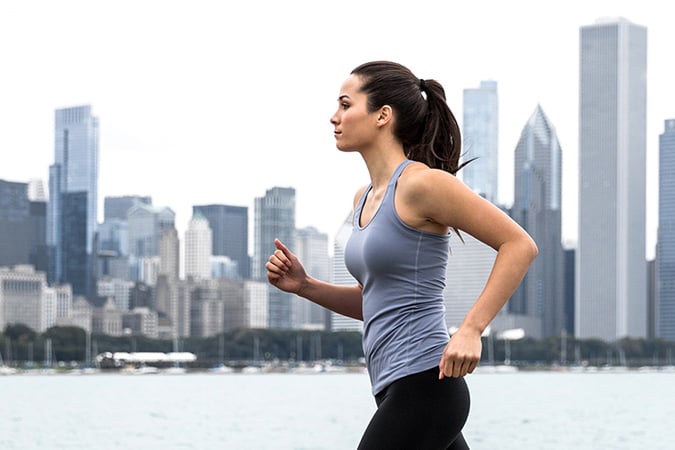 A woman wearing a grey vest top and black leggings jogs outdoors along a waterfront, with a city skyline featuring tall buildings in the background.