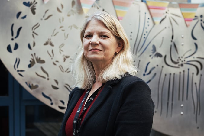 Ellen Copson, wearing a black blazer and red top stands in front of a decorative metal sculpture with abstract patterns and colourful bunting overhead.