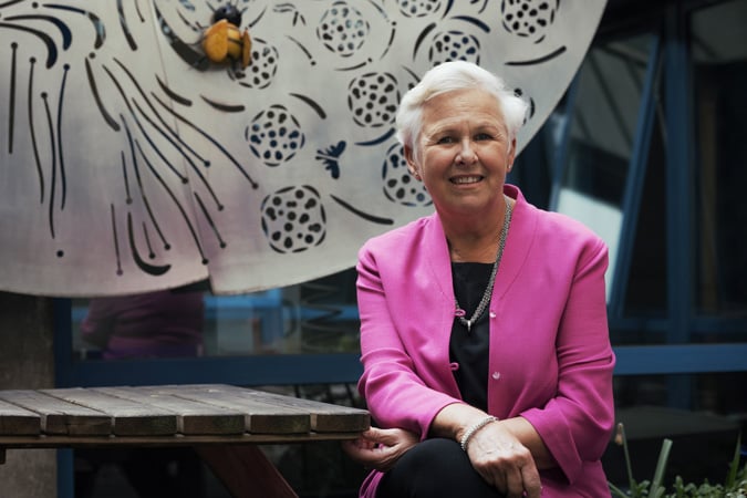 Lesley Turner, wearing a pink blazer and black outfit, sits and smiles outside beside a wooden table and large decorative metal artwork.