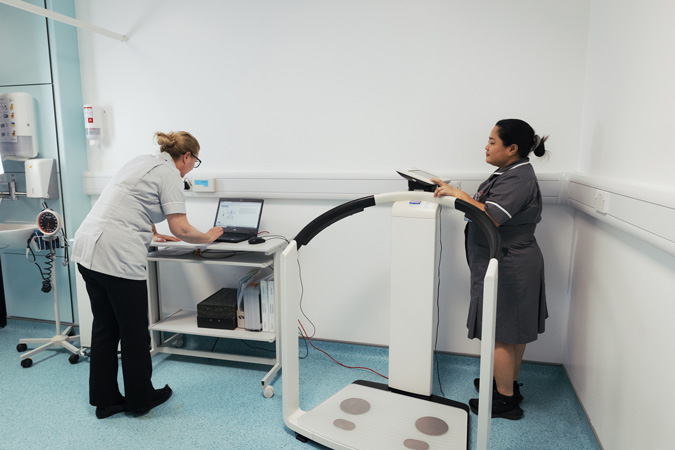 Two healthcare workers in uniforms are in a medical examination room. One is using a laptop on a mobile trolley, while the other stands on a body composition analyser scale. The room is clean and equipped with medical devices.