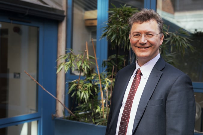 Ramsey Cutress in a suit and red patterned tie stands smiling outdoors near a blue-framed glass door and green potted plants.