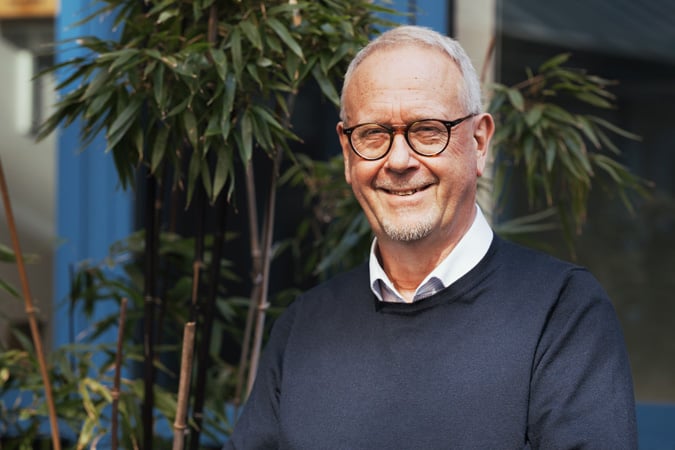 Steve Wootton, standing outdoors in front of green plants and a blue wall, wearing a navy jumper over a collared shirt.