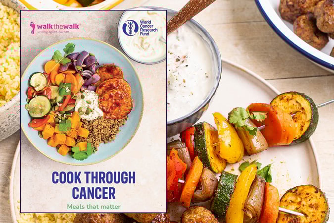 A cookbook titled Cook Through Cancer with a plate of colourful vegetables, grains, and sauce on the cover. Around it are dishes of couscous and vegetable kebabs on a wooden table.