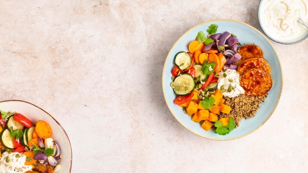 A colourful bowl of roasted vegetables, grains, two small patties, fresh herbs, seeds, and a dollop of creamy sauce, with a side bowl of yoghurt on a light, textured background.