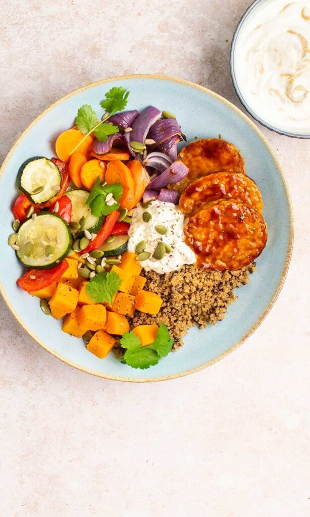 A colourful bowl with quinoa, sliced carrots, cucumber, red onion, pepper, roasted sweet potato, barbecue-glazed patties, coriander, seeds, and a dollop of creamy sauce, with extra sauce in a small bowl nearby.