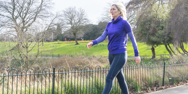 A woman in sportswear walks briskly along a park path on a bright, clear day, with green grass, trees, and a black metal fence in the background.