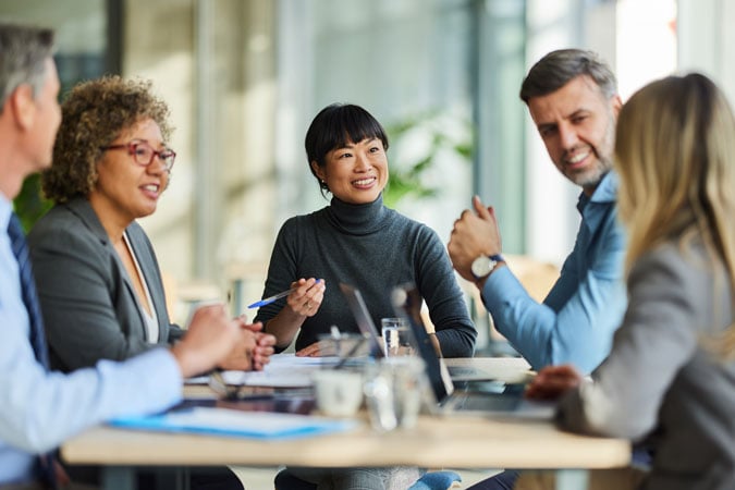 A group of five professionals sits around a table in a modern office, engaged in a discussion. They are smiling and appear to be collaborating, with documents and a laptop on the table.