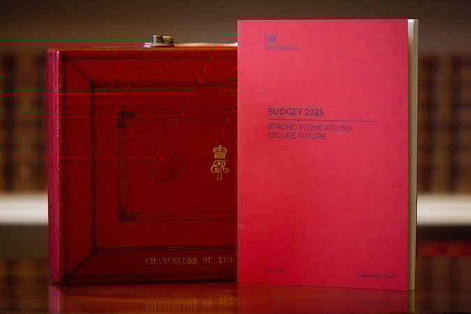 A red document titled Budget 2025: Strong Foundations, Secure Future stands upright next to a red briefcase labelled Chancellor of the Exchequer with a gold emblem and lettering.