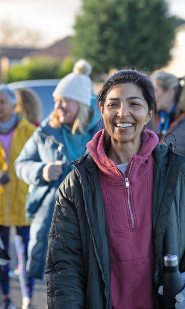 A woman in a green jacket and pink hoodie smiles outdoors, holding a water bottle. Several other people in colourful winter clothing are blurred in the background. It appears to be a cheerful group gathering or event.