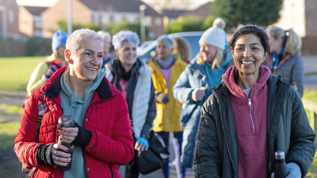 A group of women dressed in colourful jackets and winter hats smile and walk outdoors together, carrying water bottles, on a bright, sunny day in a suburban neighbourhood.