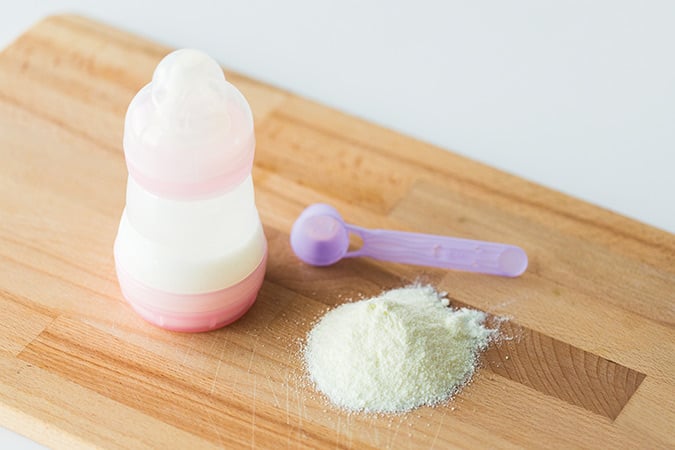A baby bottle filled with milk stands next to a small pile of powdered formula and a purple plastic scoop on a wooden surface.