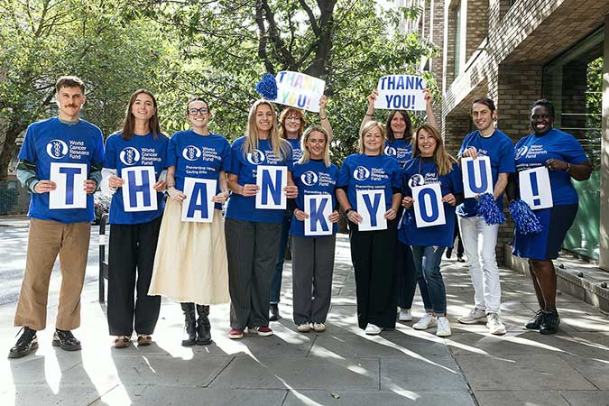 A group of people wearing matching blue shirts stand outdoors, smiling and holding large letters that spell THANK YOU! whilst others hold signs saying THANK YOU! and blue pom-poms. Trees and buildings are in the background.