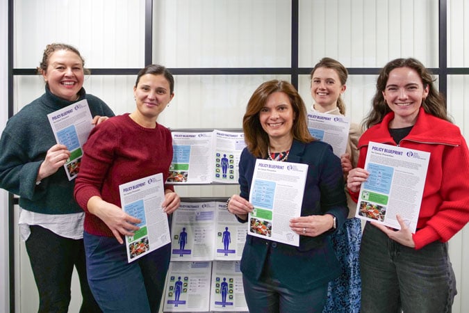 The Policy and Public Affairs team, smiling and holding printed documents titled Policy Blueprint. Behind them are more copies of these documents displayed on a glass wall. They appear to be part of a team or group presentation.