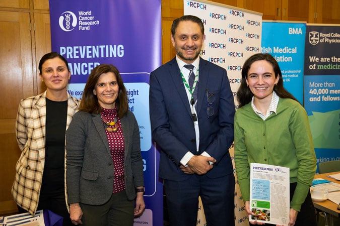Four professionally dressed people stand smiling in front of informational banners about cancer prevention and medical organisations at an indoor event. One person holds a magazine or brochure.