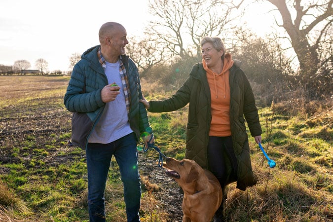 A man and woman walk together outdoors on a grassy path with a brown dog on a lead. Both are smiling and dressed warmly. The sun is shining and trees are visible in the background.