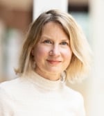 A woman with light blonde hair and fair skin, wearing a cream polo neck jumper, stands indoors in soft natural light with a blurred background. She smiles gently at the camera, embodying the warm spirit of CUP Global.