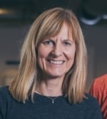A woman with straight blonde hair, wearing a dark top and a necklace, smiles at the camera in an indoor setting.