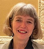 A woman with short, grey hair and a fringe smiles at the camera. She is wearing a dark top and is standing in front of a light, ornate wall with decorative moulding.